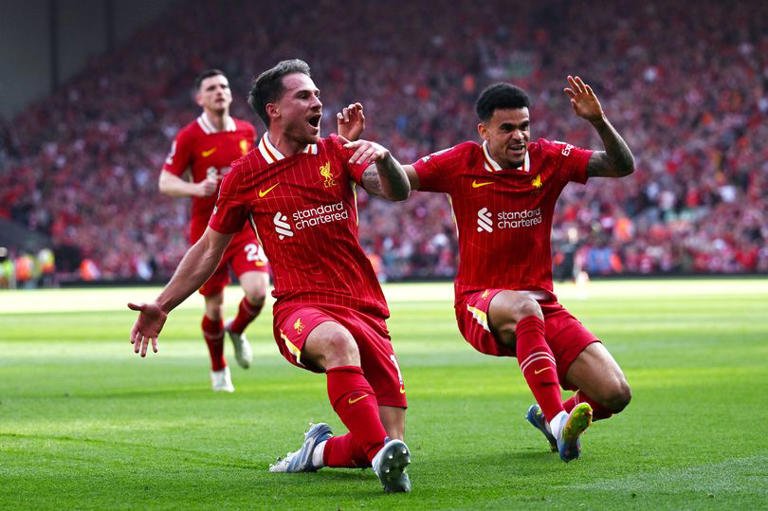 Soccer players in red uniforms celebrating a goal on the field, with a stadium crowd in the background.