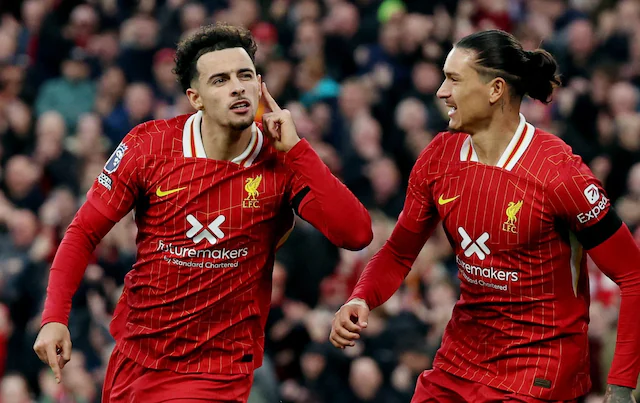 Two soccer players in red and white Liverpool uniforms celebrating on the field. One is gesturing to his ear and the other is smiling. A crowd is visible in the background.