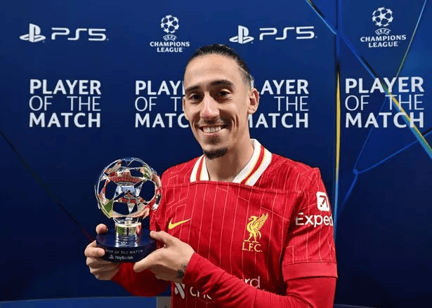 Soccer player in red Liverpool FC jersey holding "Player of the Match" trophy in front of Champions League backdrop.