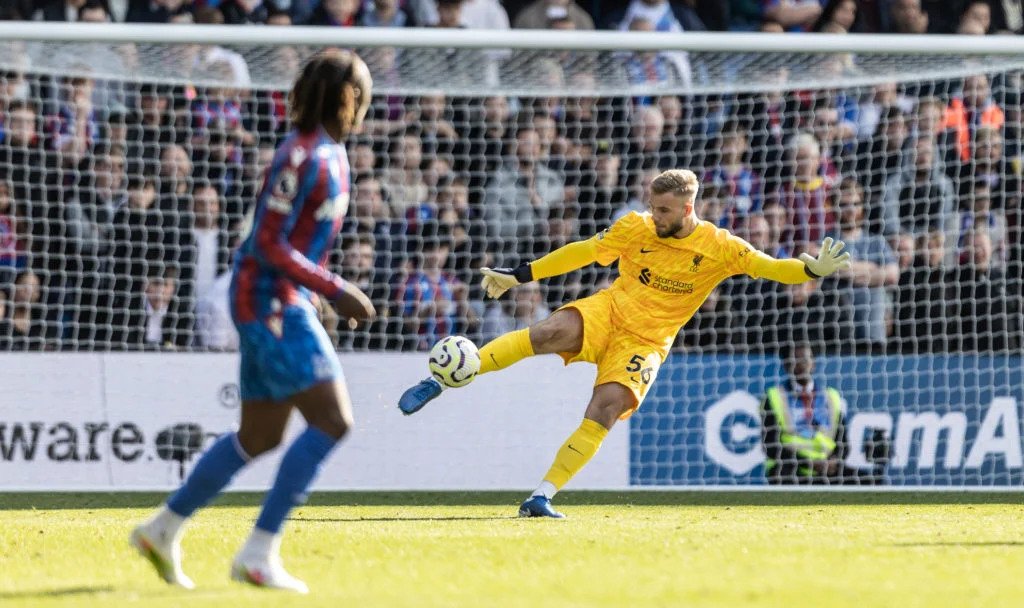Soccer goalkeeper in yellow kit kicking the ball during a match, with an opposing player in blue and red in the foreground, set in a stadium with a crowd in the background.