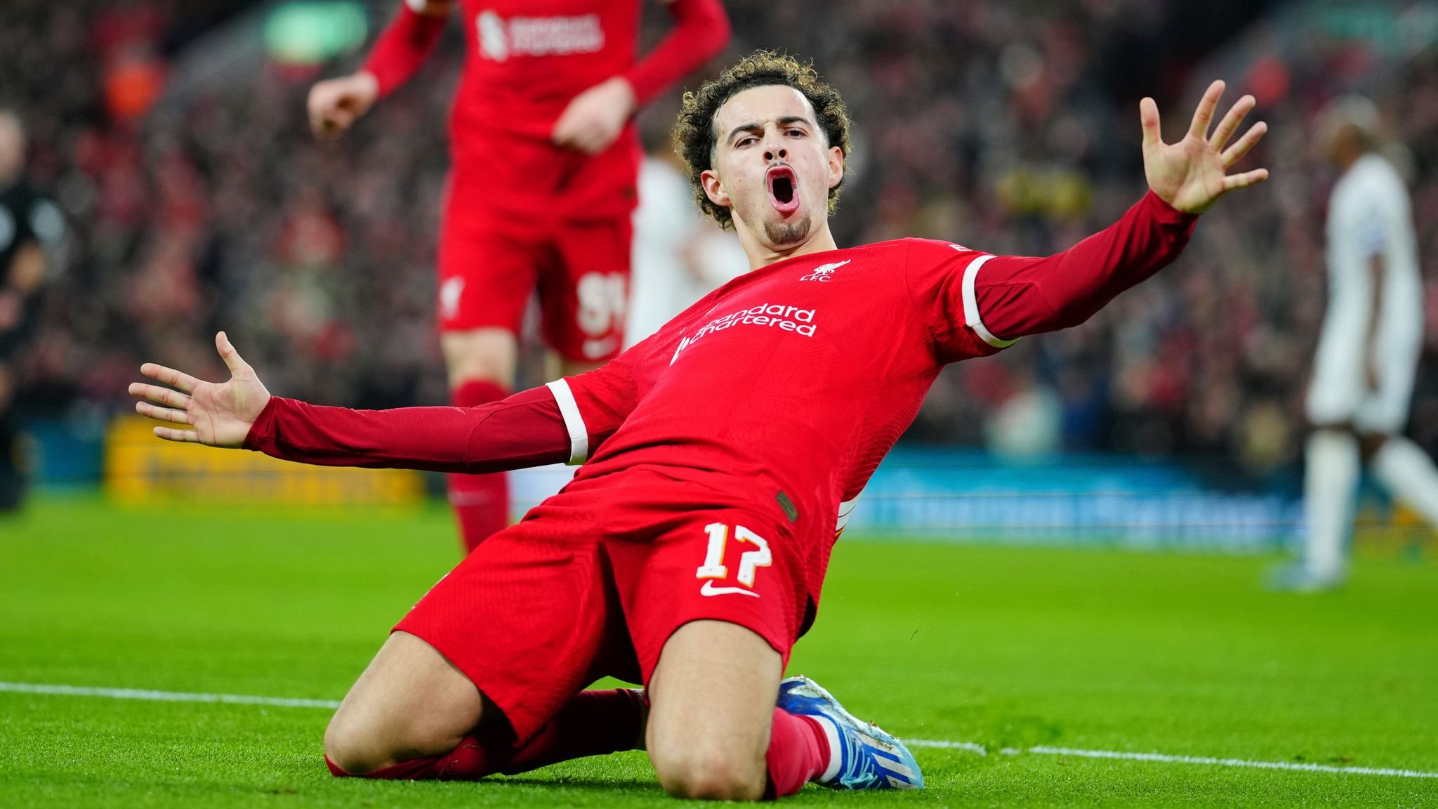 Soccer player in red uniform celebrating a goal by sliding on knees on the field, arms spread wide.