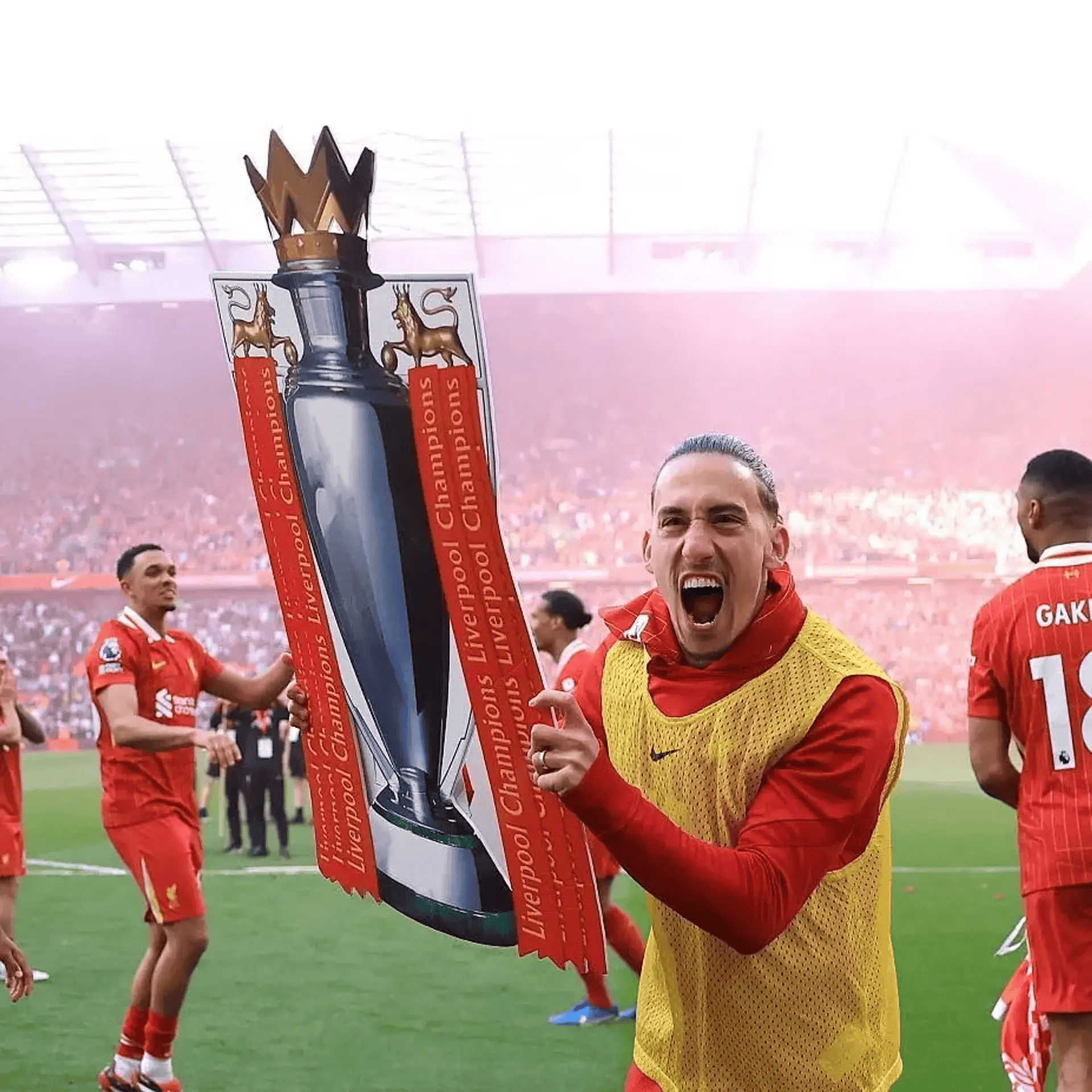A soccer player celebrating with a cardboard cutout of a trophy on a football field, surrounded by teammates in red jerseys. The trophy cutout has the words "Liverpool Champions."