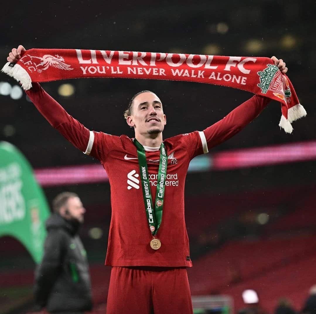 Soccer player in a red Liverpool FC jersey holds a "You'll Never Walk Alone" scarf over their head, wearing a medal and standing on a field, celebrating.