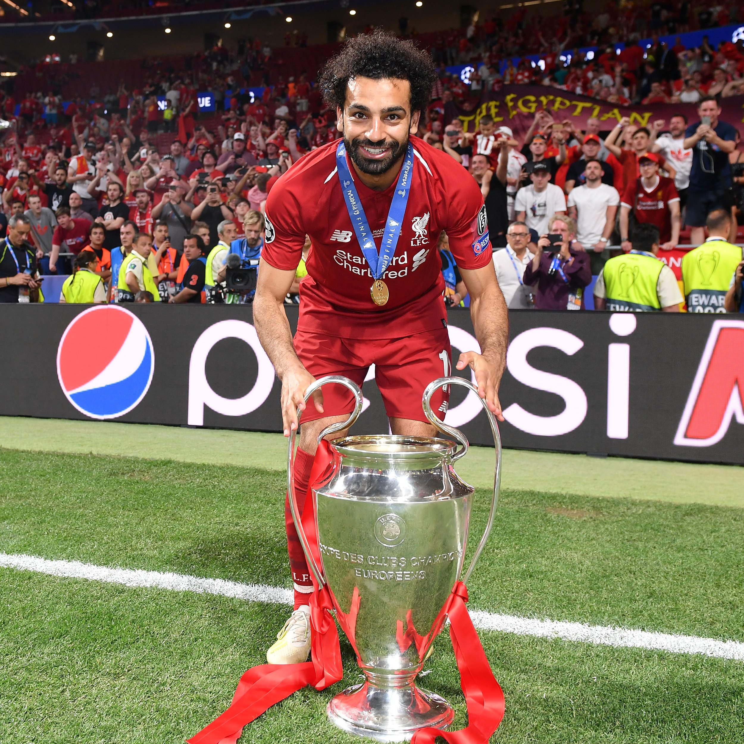 A soccer player in a red jersey holding a large trophy on a football field, surrounded by jubilant fans. The trophy has red ribbons attached, and the player is wearing a medal.