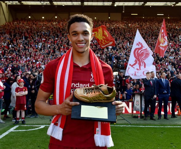 Soccer player in red jersey holding a golden shoe trophy with a cheering crowd and Liverpool FC flags in the background.