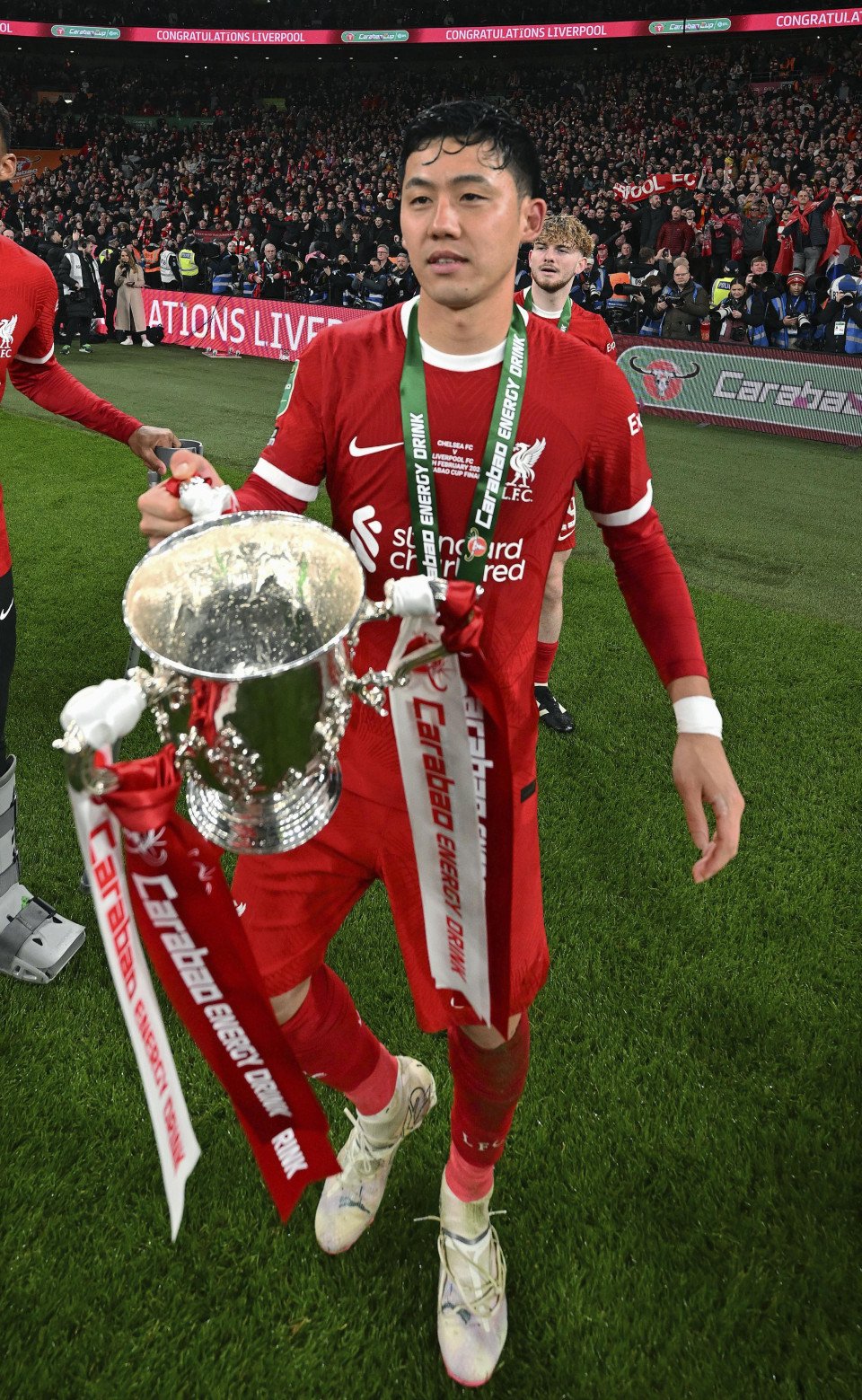 Soccer player in red uniform holding a trophy with ribbons, standing on a grass field with spectators in the background.