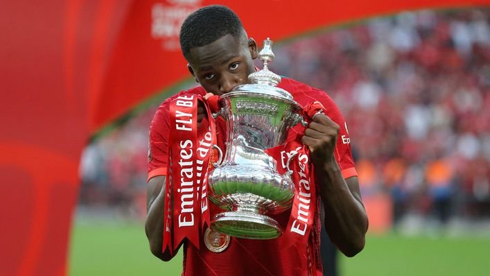 Soccer player in red jersey kissing FA Cup trophy with red ribbons on a sports field.