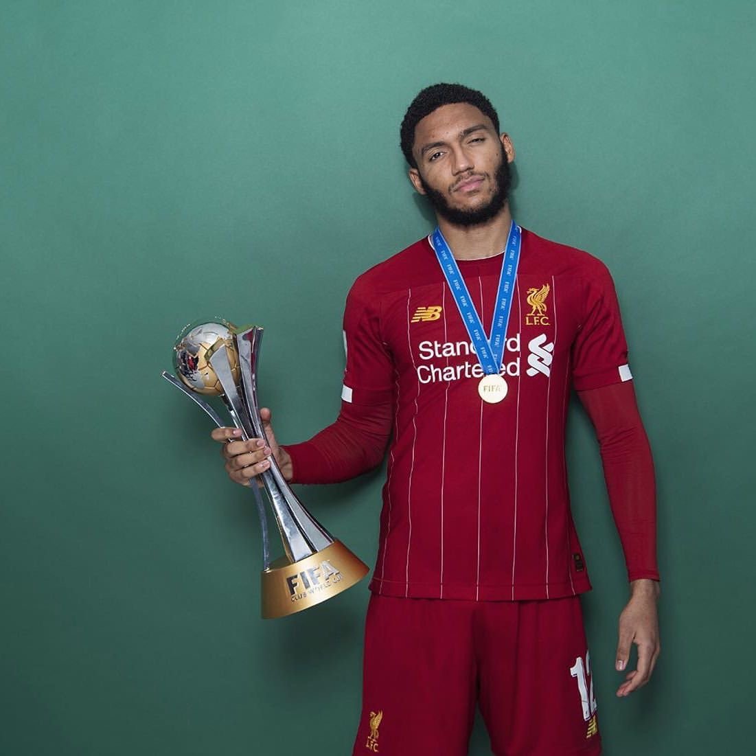 Soccer player in a red Liverpool FC jersey holding a FIFA Club World Cup trophy with a medal around his neck against a green background.