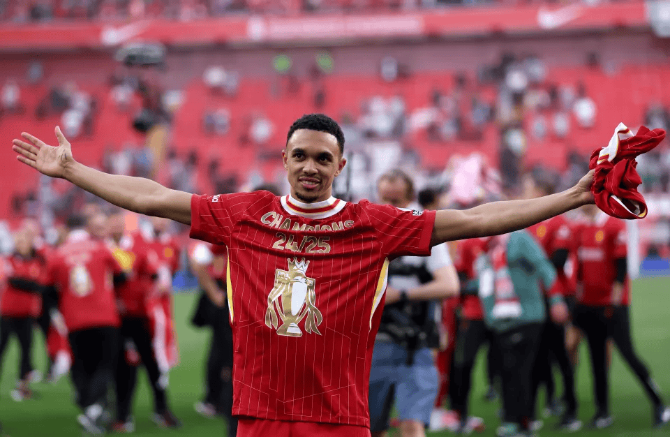 Soccer player celebrating with arms outstretched, wearing a red jersey with 'Champions 24/25' printed, on a sports field with blurred crowd in the background.