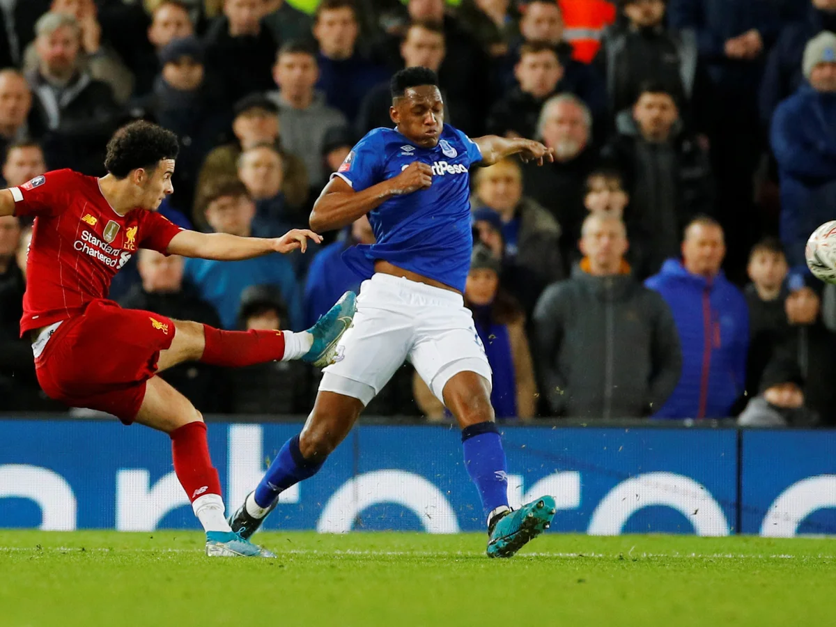Soccer players from Liverpool and Everton in action during a match, with one player in red shooting at the goal and another in blue defending.