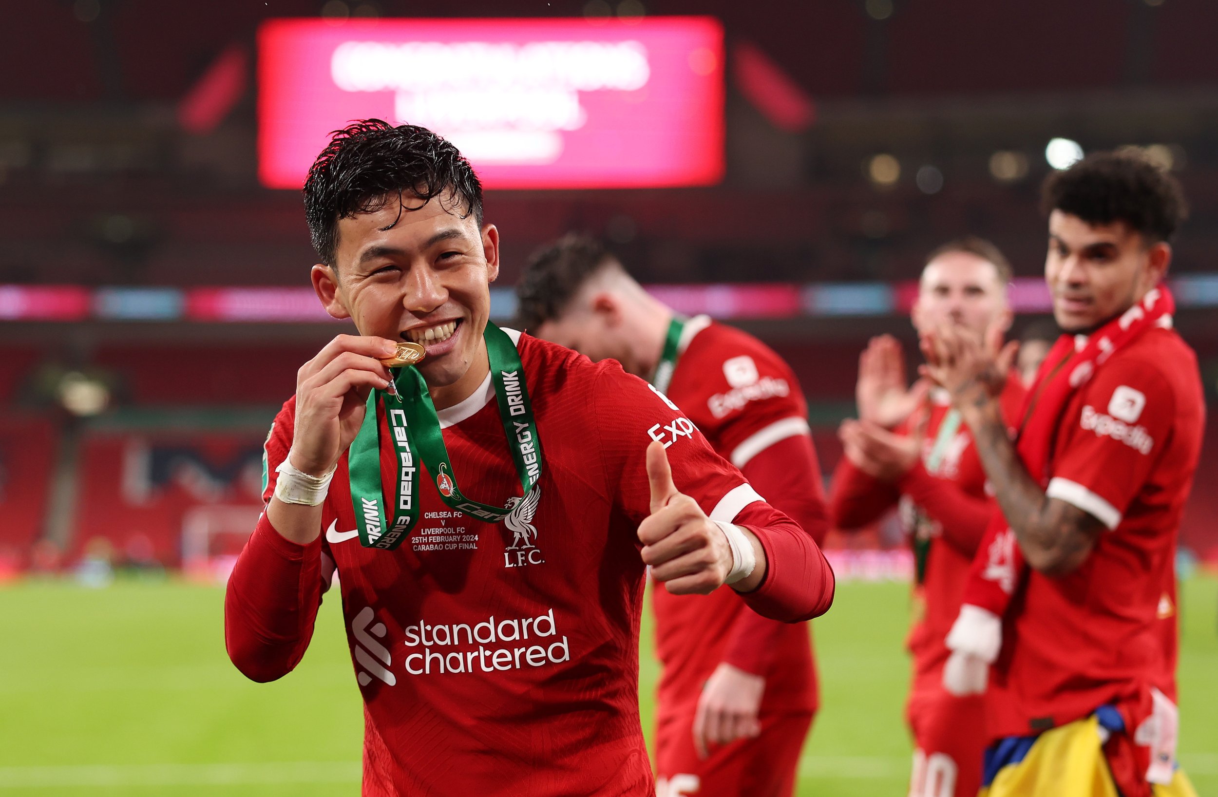 Soccer player in red Liverpool jersey holding a medal, biting it, and giving a thumbs up after a match victory.