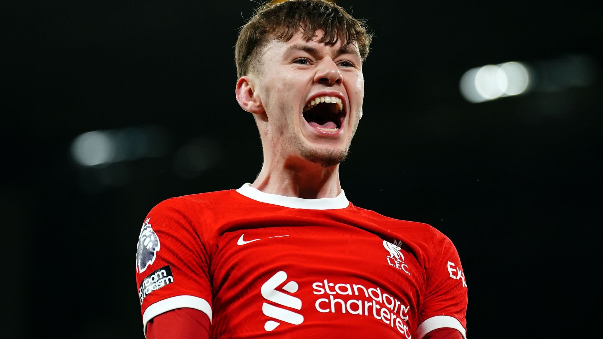 Soccer player in red uniform celebrating with open mouth, Premier League badge visible on sleeve.