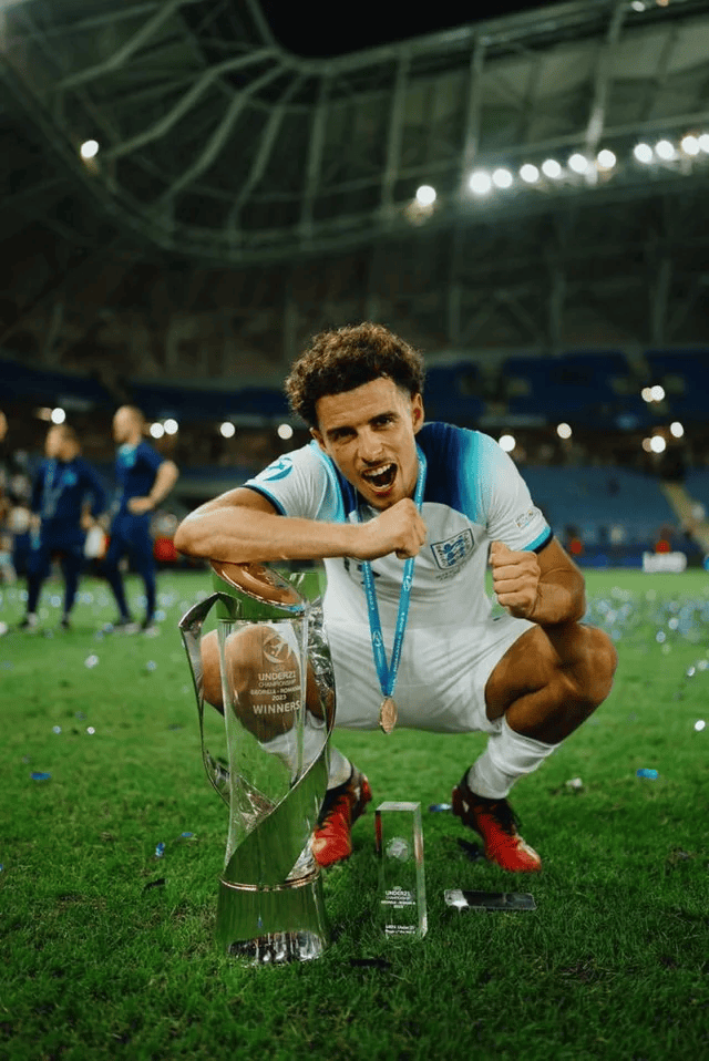 Soccer player celebrating with trophies on a field inside a stadium. The player is wearing a white and blue jersey and holding a medal, with a large trophy labeled 'Under-21 Winners' in front. Background shows stadium seating and scattered confetti.