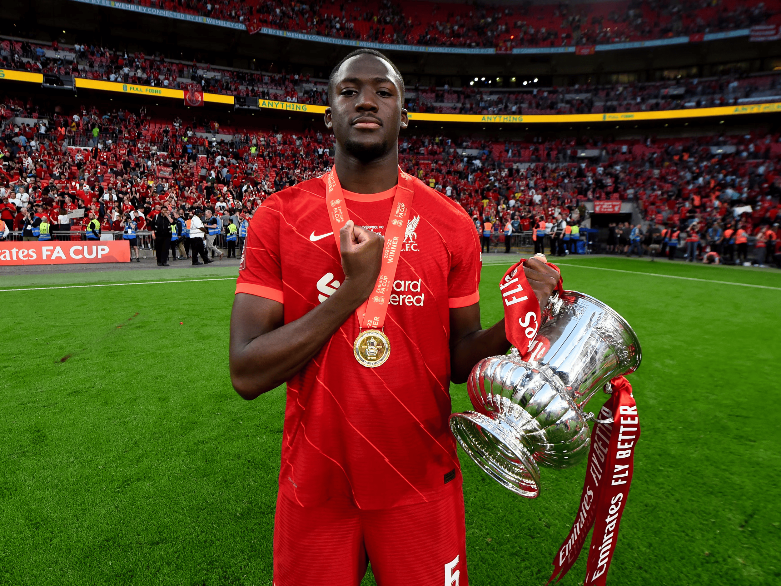Soccer player in red jersey holding an FA Cup trophy on a football field with a crowd in the background.