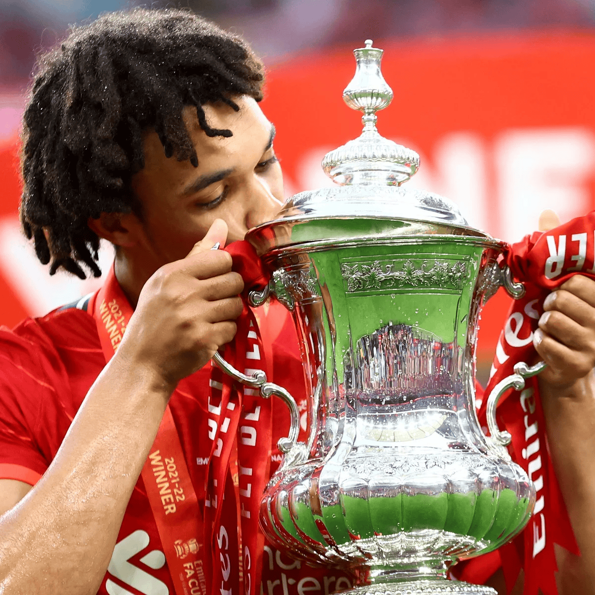 A soccer player in a red jersey kissing a large, ornate silver trophy with red ribbons attached, likely after winning a football match. The player is wearing a winner's medal around his neck.
