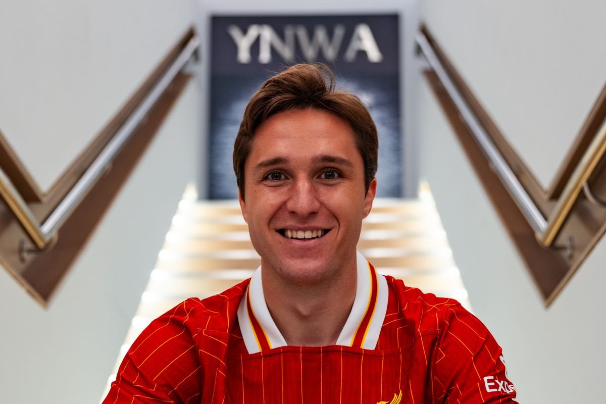 A person smiling, wearing a red sports jersey with yellow accents, in front of a staircase with the letters "YNWA" in the background.