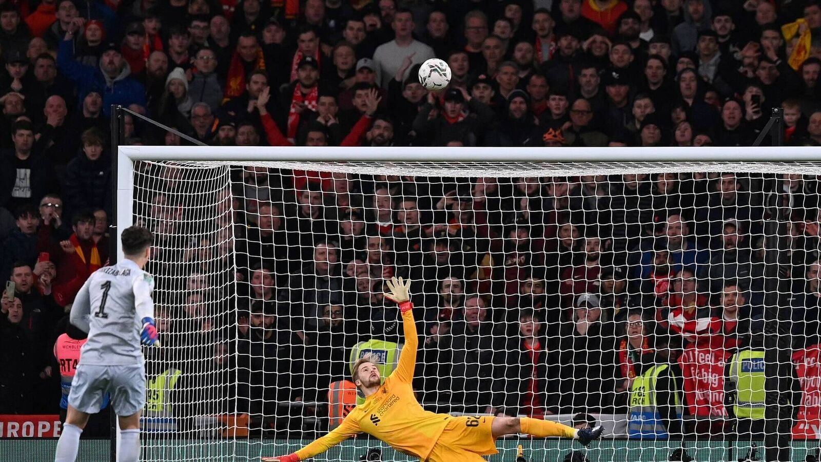 Soccer goalkeeper in orange attempting to save a penalty kick while the ball goes over the net, with a crowd watching intensely in the background.