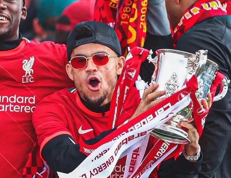Person wearing a red Liverpool FC jersey and sunglasses, holding a silver trophy wrapped in red and white ribbons, celebrating with an open mouth.