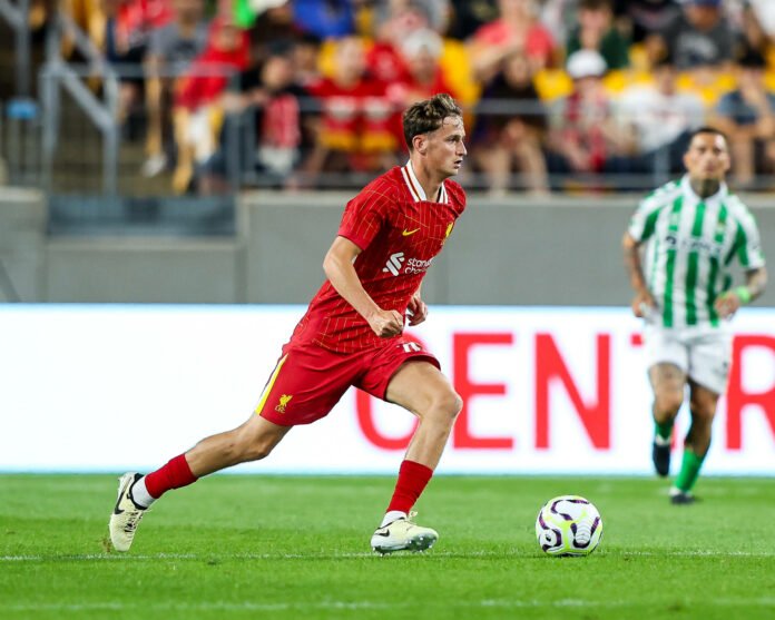 Soccer player in red jersey dribbling a ball during a match, with another player in a green-and-white striped jersey in the background, on a grassy field.