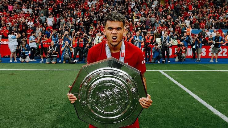 A soccer player in a red uniform holding a large silver trophy on a field, with a crowd in the background celebrating.