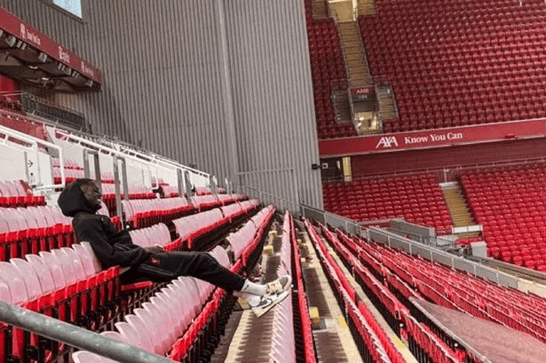 Person sitting alone in a stadium, wearing a hoodie and sneakers, surrounded by empty red seats.