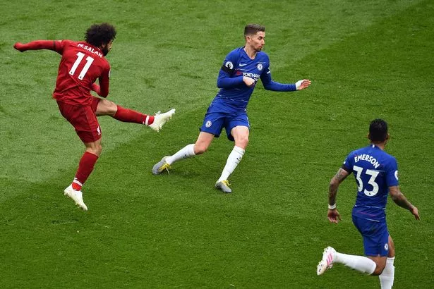 A soccer player in a red jersey kicking a ball during a match, while two opposing players in blue jerseys move towards him on a grass field.