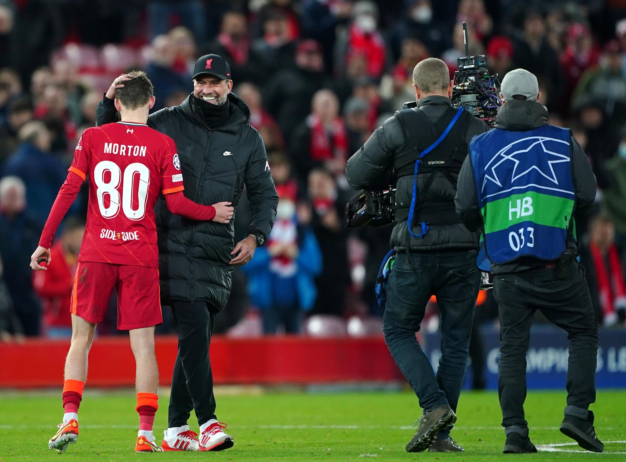 Soccer player in red jersey with number 80 and "MORTON" on the back, interacting with a man in a black jacket and baseball cap on a field, with camera crew filming them.