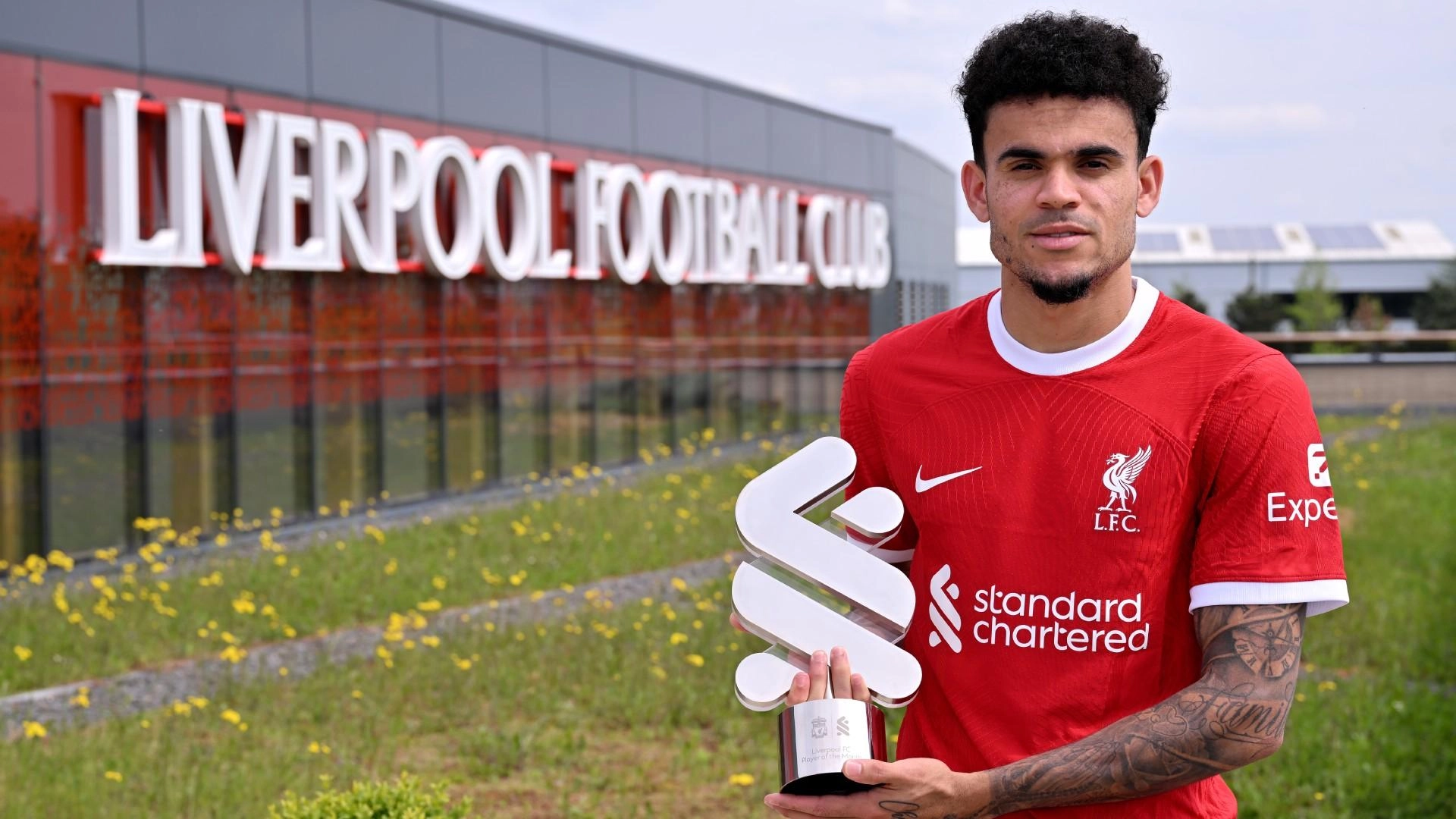 Soccer player in a red Liverpool FC jersey holding a trophy in front of the Liverpool Football Club building.