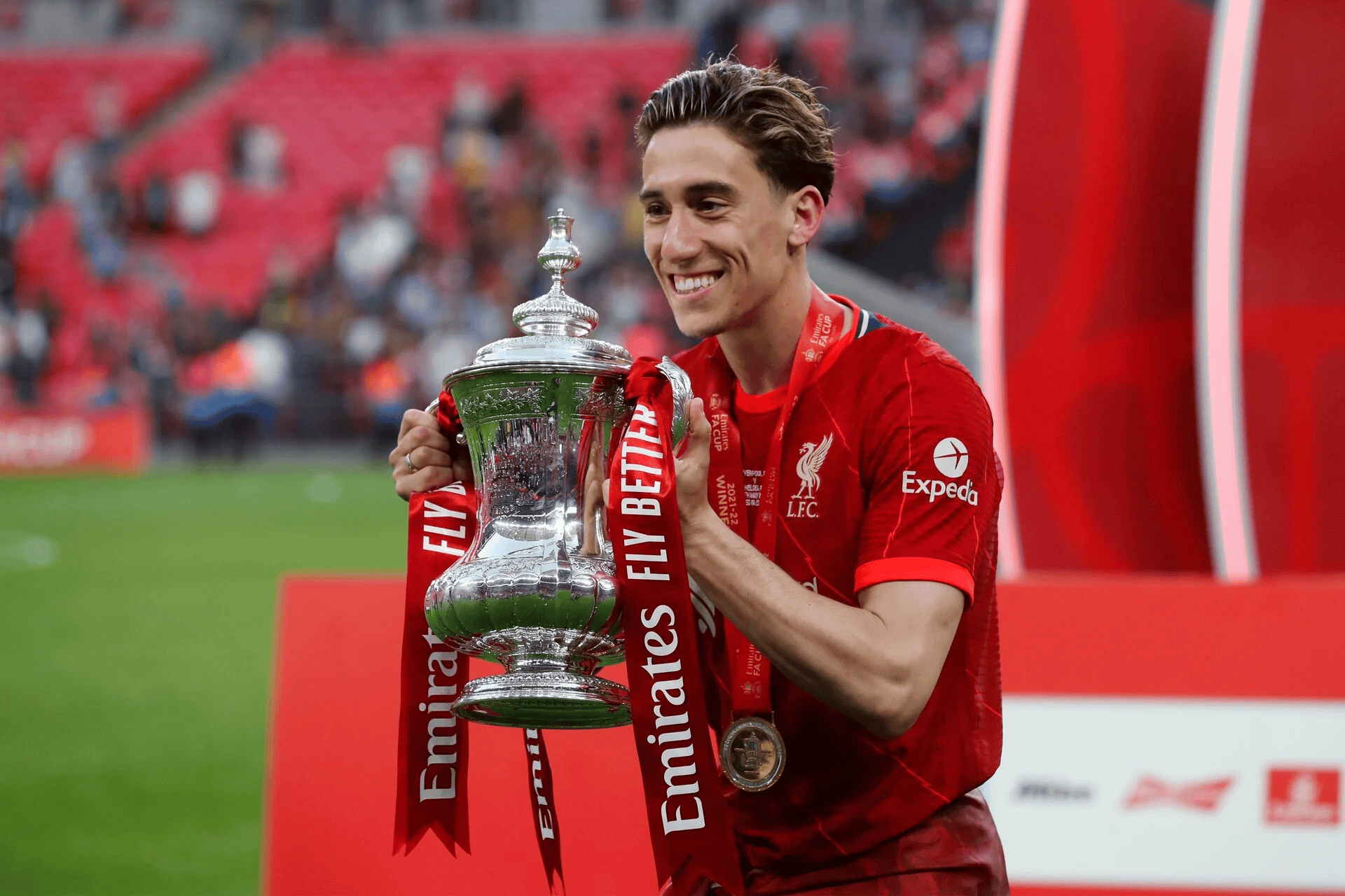 Soccer player in red Liverpool jersey holding the FA Cup trophy on a field, with red Emirates ribbons on the trophy.