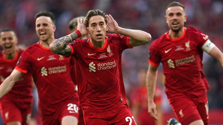 Soccer players from Liverpool FC celebrating on the field, wearing red uniforms with sponsors' logos, one player gesturing excitedly with his hands near his ears.
