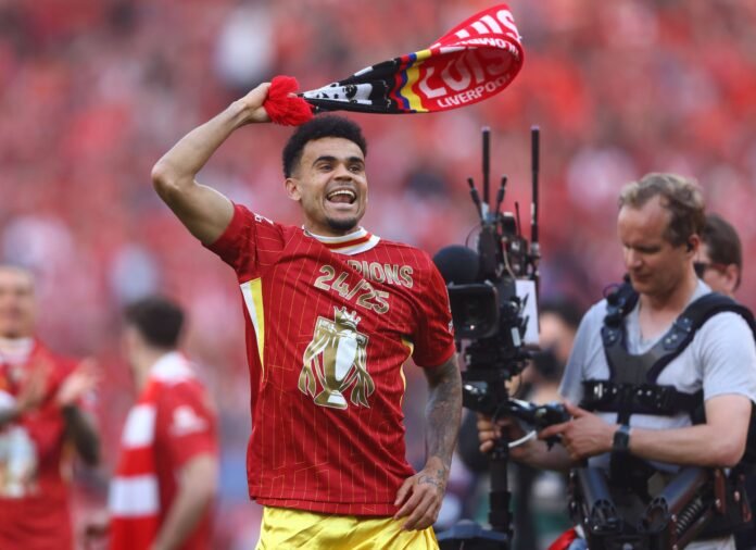Soccer player celebrating with a Liverpool scarf and wearing a red "Champions" jersey, on a field with a cameraman nearby.