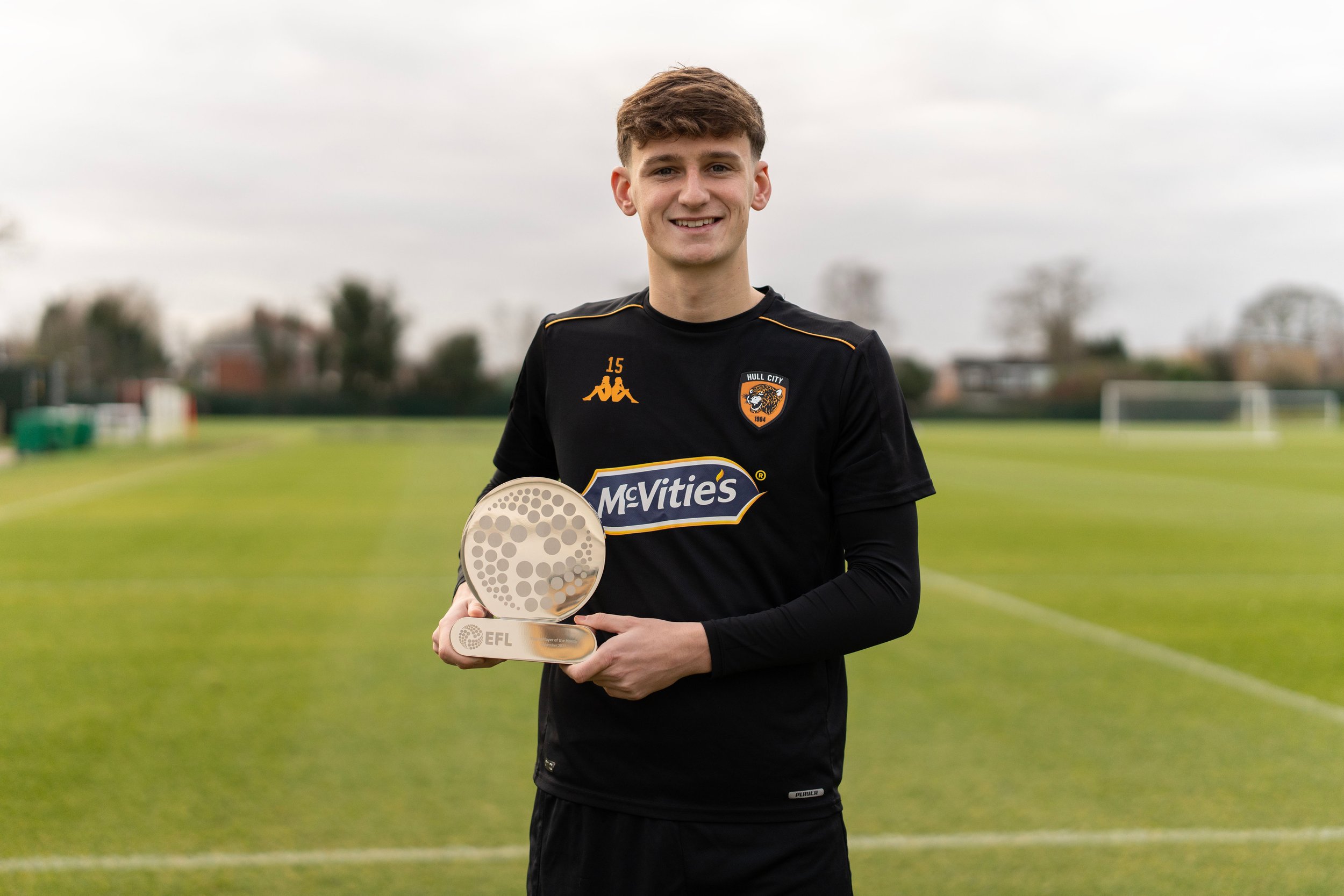 A soccer player wearing a black Hull City jersey holds an EFL award on a field.