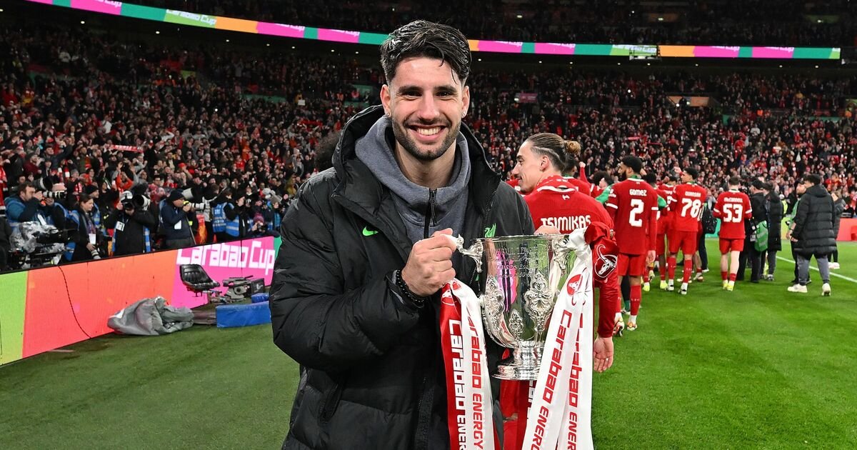 A person holding a Carabao Cup trophy on a soccer field, surrounded by cheering fans and team players. The atmosphere is celebratory, with other players wearing red jerseys in the background.