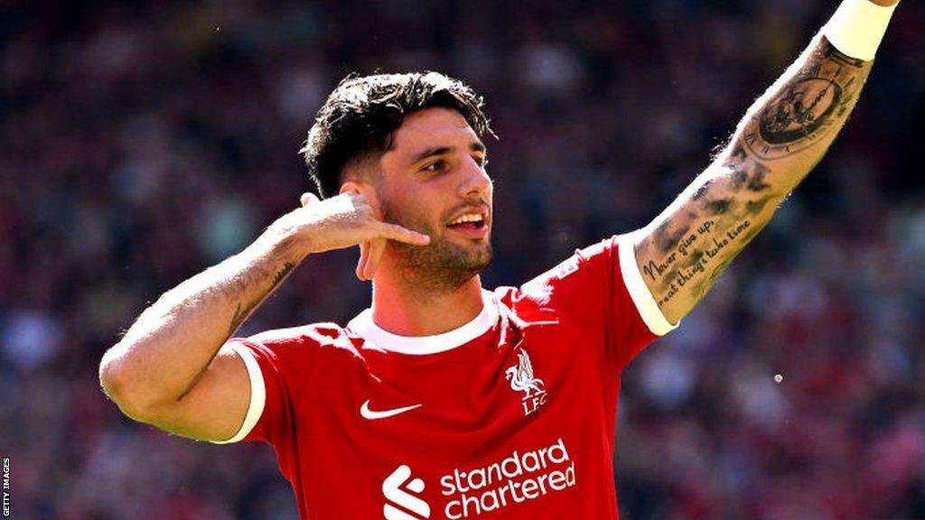 Soccer player in a red jersey with tattoos on his arm, making a phone gesture with his hand.