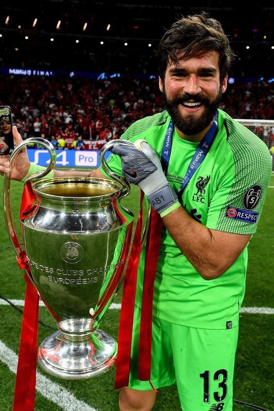Soccer player in green goalkeeper uniform holding UEFA Champions League trophy with red ribbons, smiling on the field.