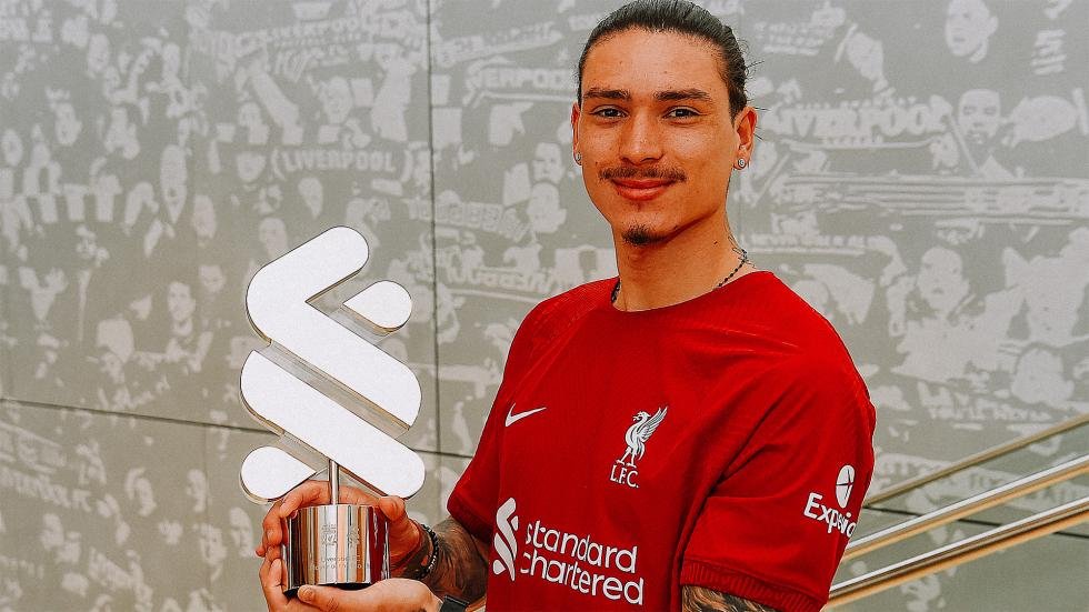 A person in a red Liverpool FC jersey holding a trophy shaped with the Standard Chartered logo, standing against a wall with images of crowds.