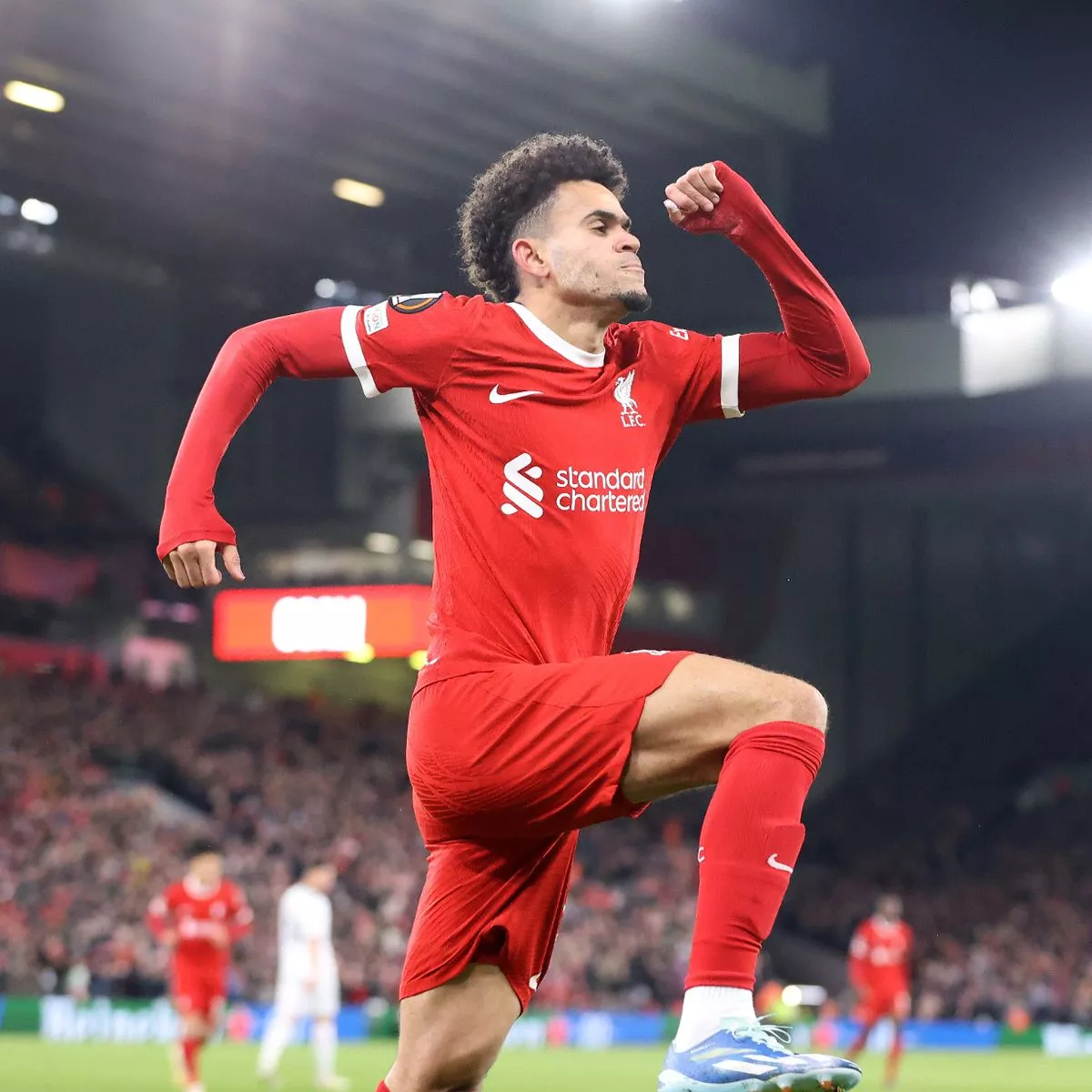 Soccer player in red jersey celebrating a goal on the field, with a stadium crowd in the background.