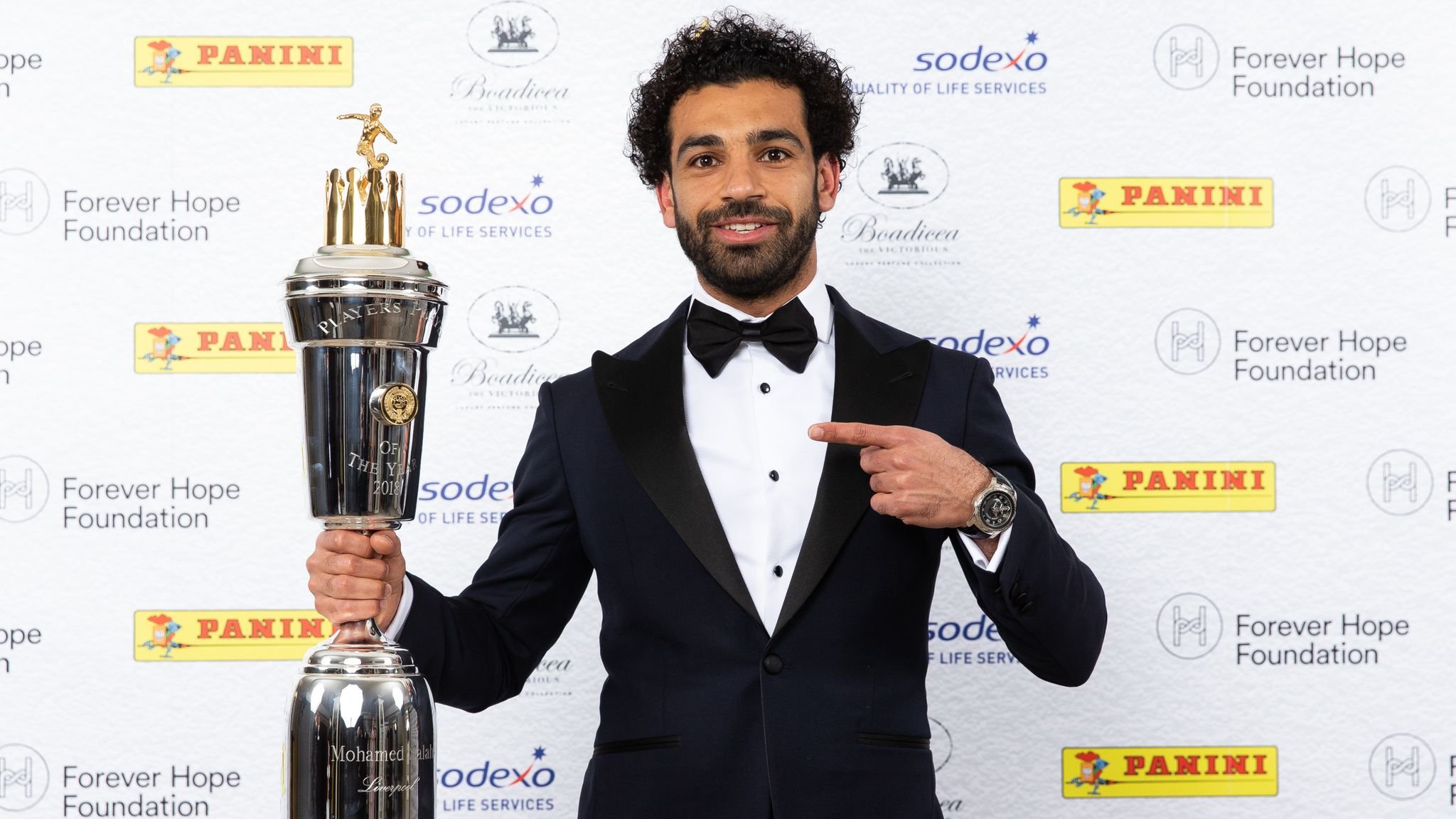 Man in tuxedo holding a sports trophy in front of a branded backdrop.