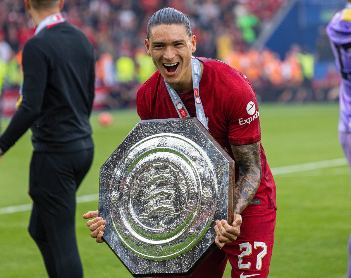Soccer player in a red jersey holding a large silver shield trophy on a soccer field after a match.