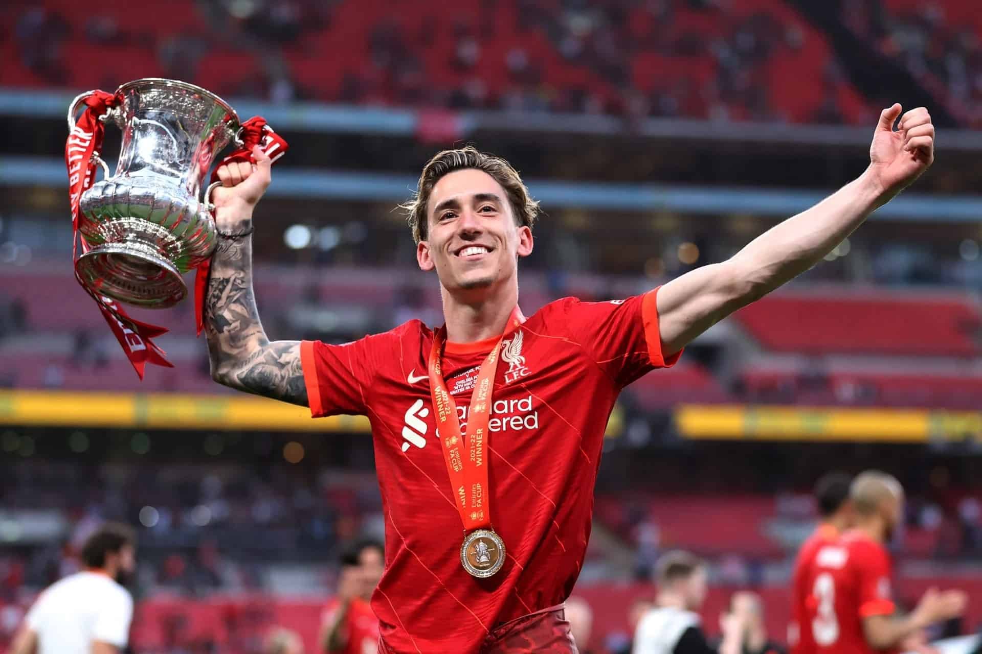 Soccer player in red jersey holding a trophy with a medal around his neck, celebrating victory in a stadium.