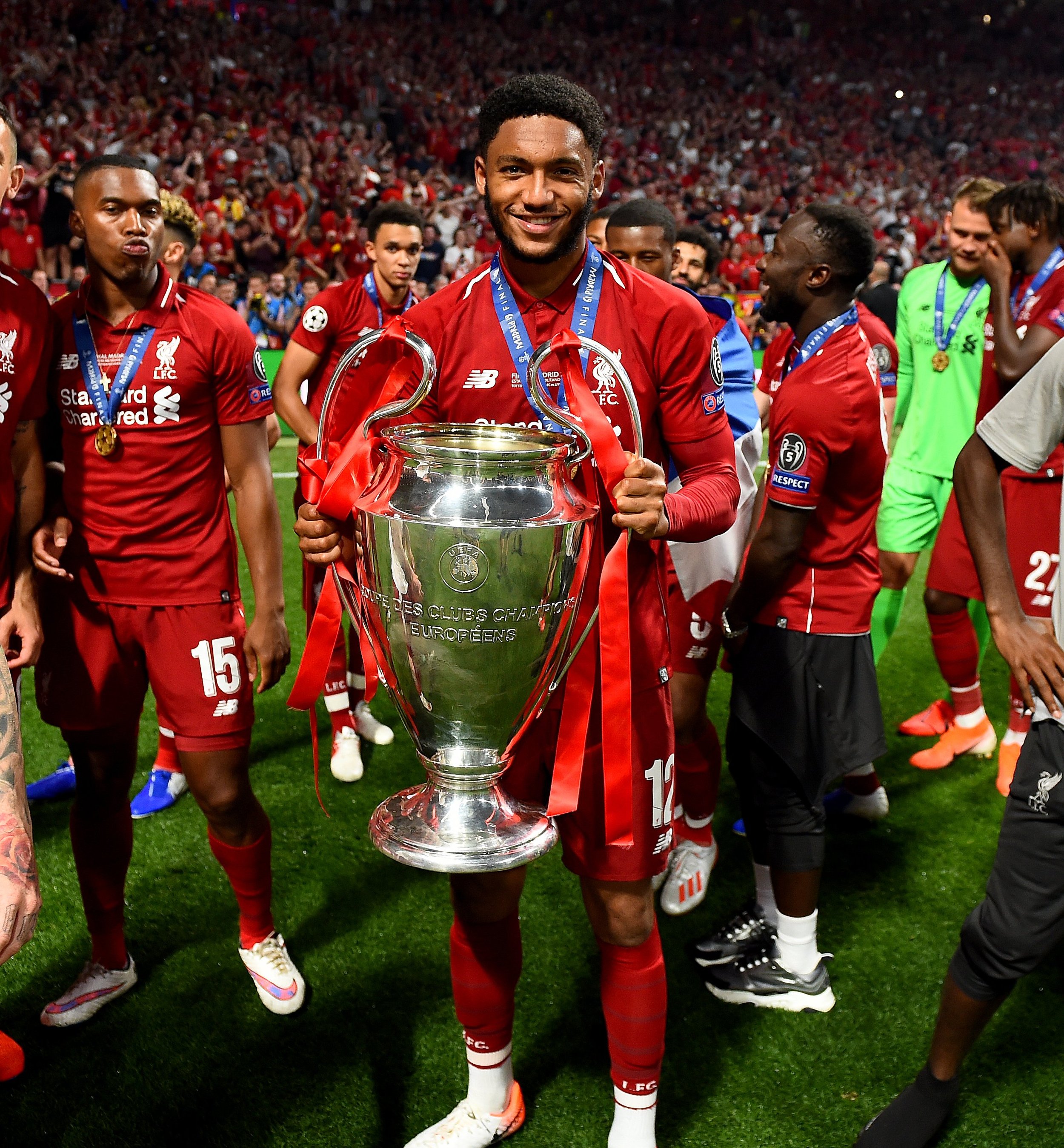 Soccer player in red jersey holding a large silver trophy with teammates on a field, celebrating.