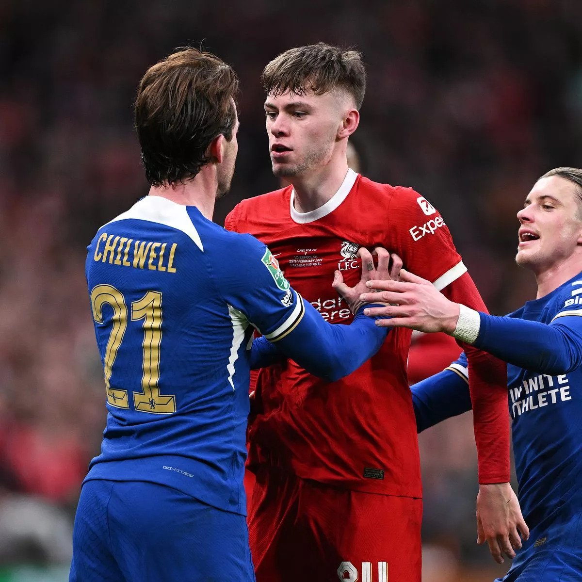 Soccer players from two different teams confront each other on the field. The player in red is being held back by two players in blue jerseys, one of whom has "Chilwell" and the number 21 on his back.