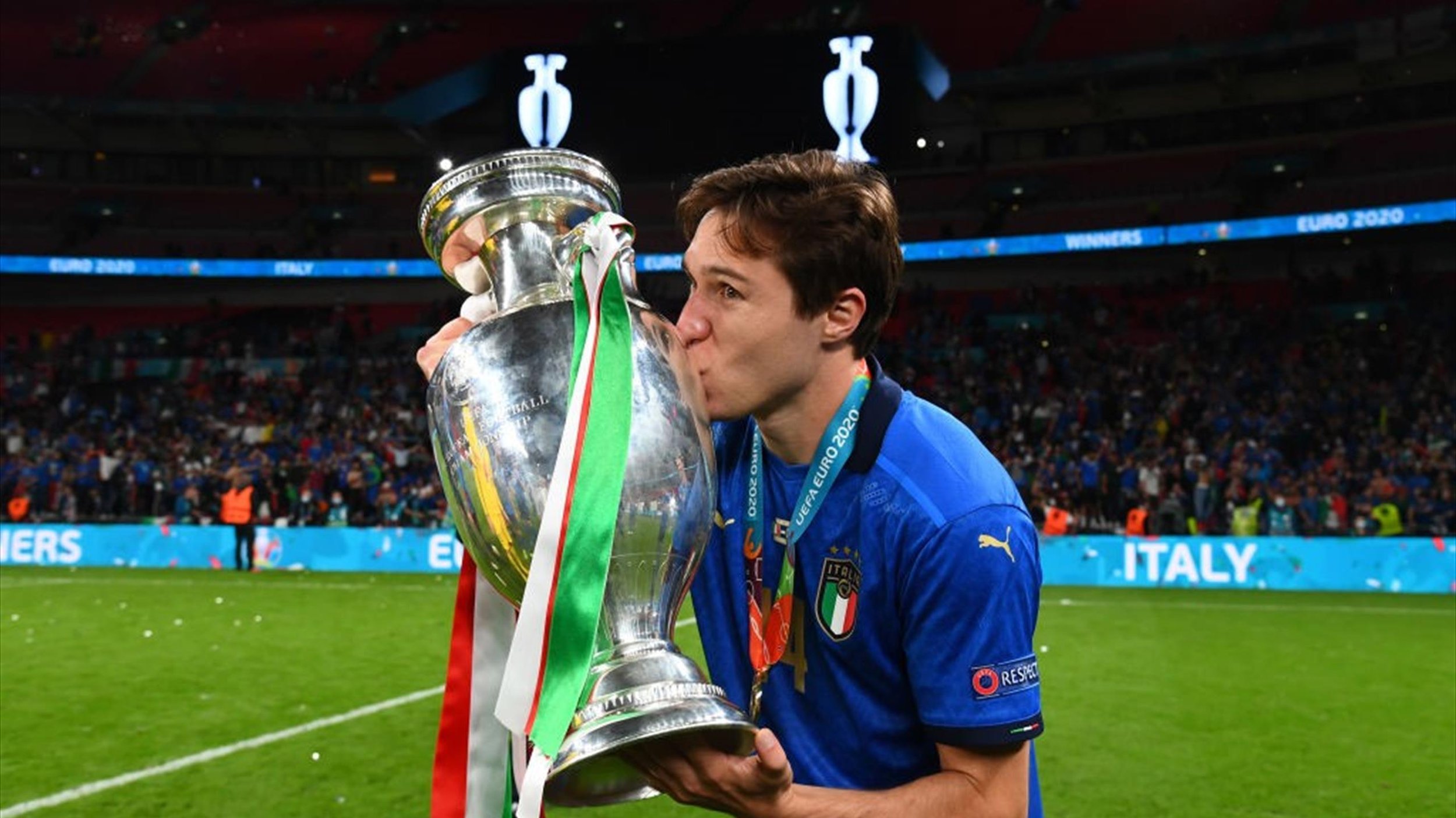 Soccer player in Italy's blue jersey kissing the UEFA Euro 2020 trophy on a field with a crowd in the background.