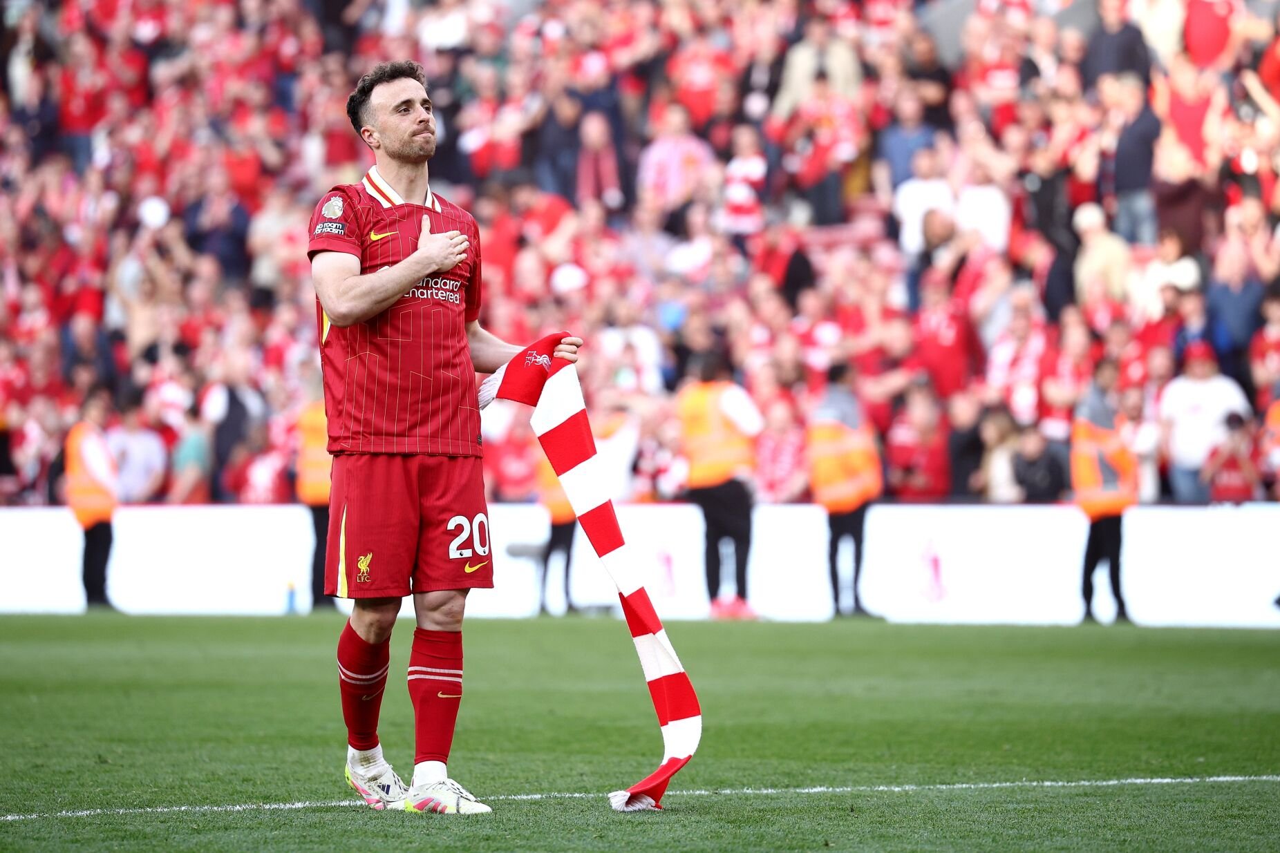 Soccer player in red Liverpool jersey holding a red and white scarf on a field, surrounded by fans.