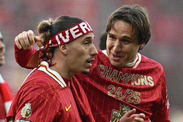 Two soccer players in red jerseys celebrate, one with a headband. Jerseys feature 'Champions 24/25' and a Premier League patch.