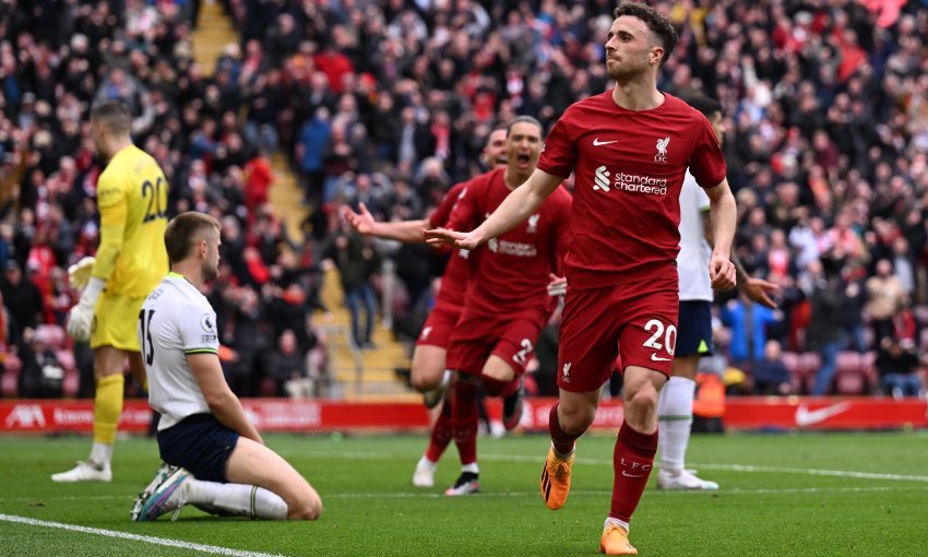 Soccer player in red jersey celebrating goal, surrounded by teammates and opponents, with a crowd in the background.