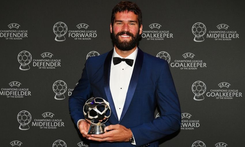 A man in a suit holding a UEFA Champions League Goalkeeper of the Season trophy, standing against a backdrop with award logos.