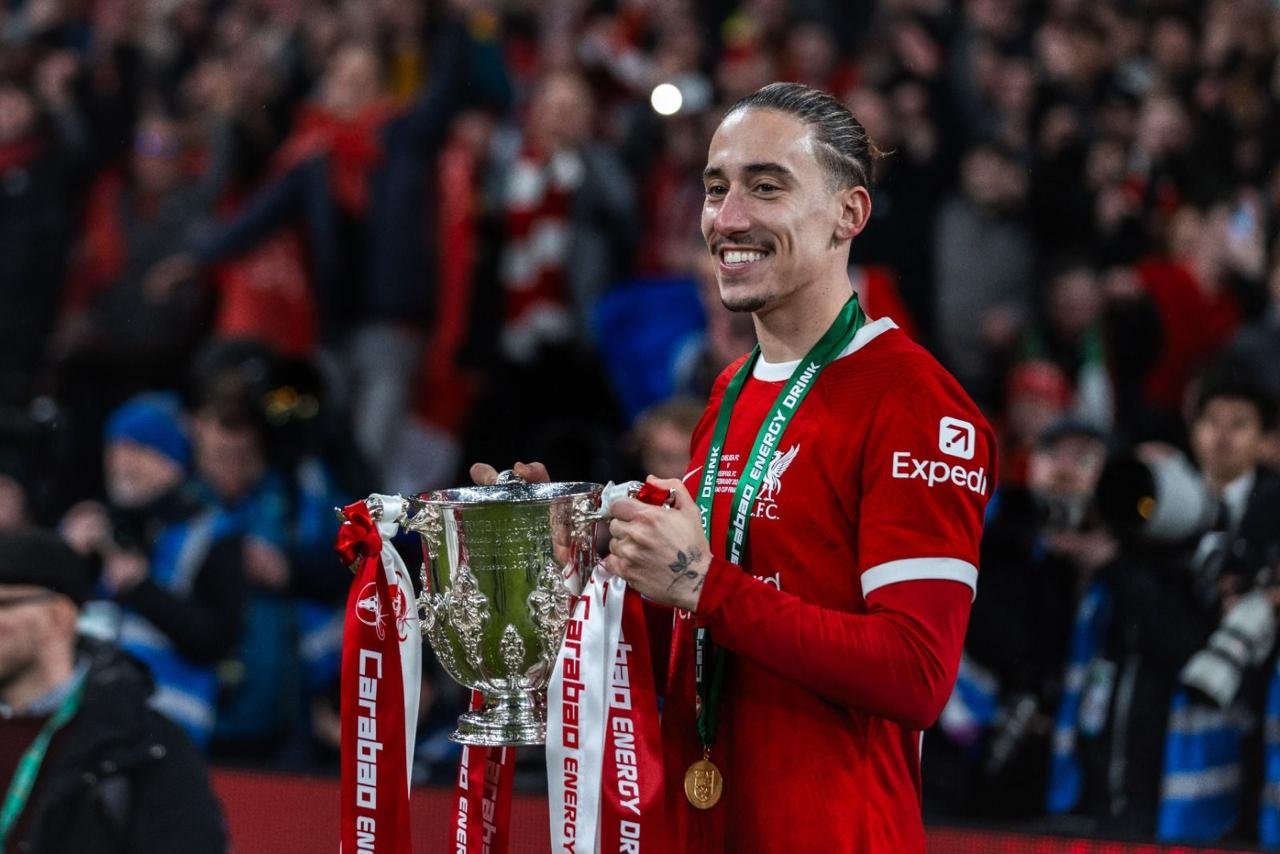 Soccer player in red jersey holding Carabao Cup trophy on field with fans in background.