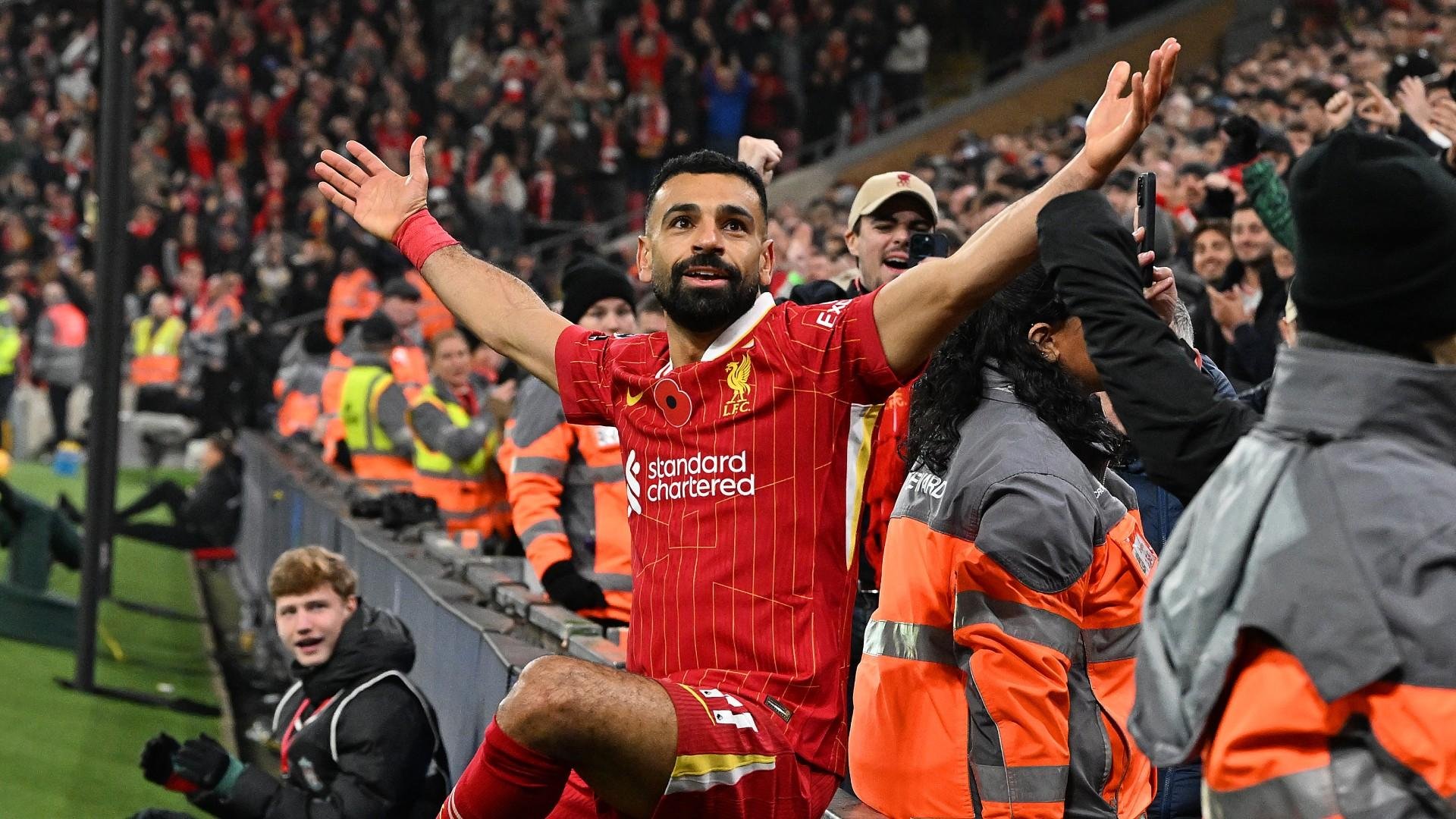 Soccer player in red Liverpool jersey celebrating with arms outstretched, surrounded by fans and security personnel in an arena.
