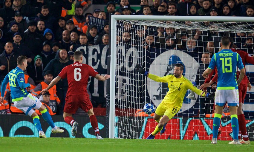 Soccer match action with a player shooting at goal, a goalkeeper in yellow attempting to block the shot, and defenders nearby. Stadium crowd in the background.